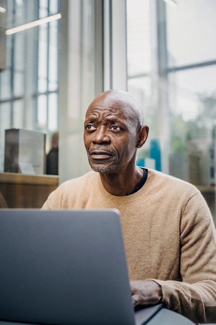 Mature African American male looking away thoughtfully while typing on laptop keyboard in sidewalk cafe